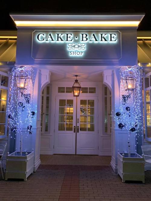 A charming cake shop entrance adorned with fairy lights and decorative trees, illuminated at night.