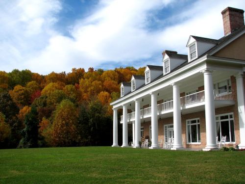 A grand mansion with white columns overlooks a vibrant autumn landscape of colorful trees under a blue sky.