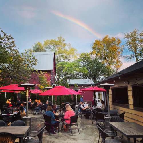 Outdoor dining area with red umbrellas, surrounded by trees, under a sky with a rainbow.