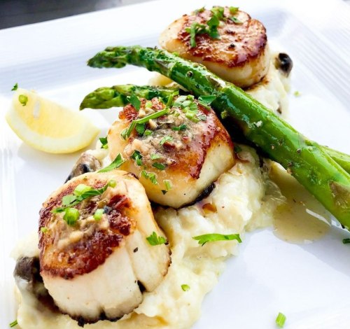 Three seared scallops on creamy mashed potatoes, garnished with asparagus and lemon wedges, topped with fresh herbs.