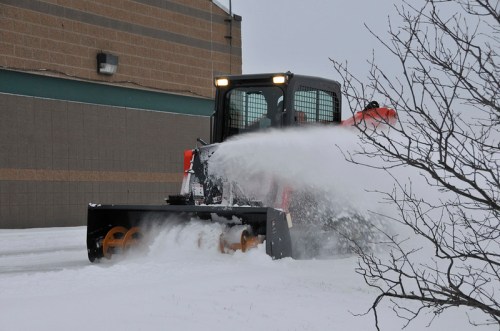 A snowplow clears a snowy parking lot near a building on a cloudy day.