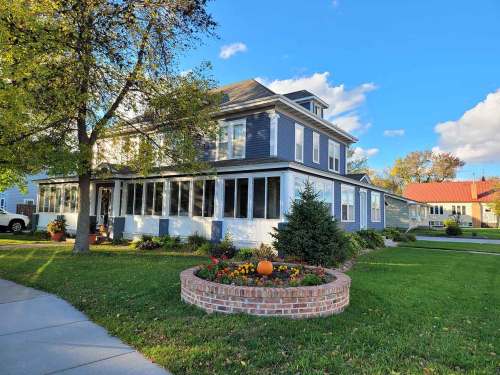 A charming two-story house with a wraparound porch, surrounded by colorful flowers and a small pumpkin in the garden.