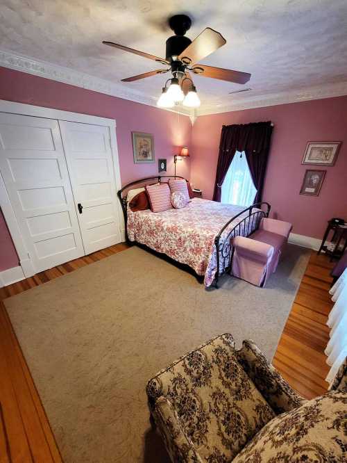 Cozy bedroom with pink walls, a floral bedspread, a ceiling fan, and vintage decor, featuring a chair and a small bench.