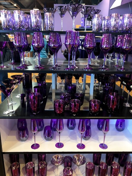 A display of various purple glassware, including wine glasses, martini glasses, and tumblers, arranged on shelves.