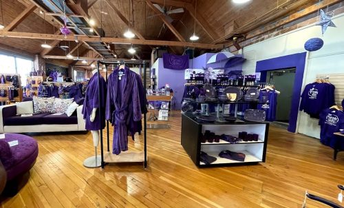 A retail store interior featuring purple merchandise, including robes, clothing, and home decor, with wooden floors and high ceilings.