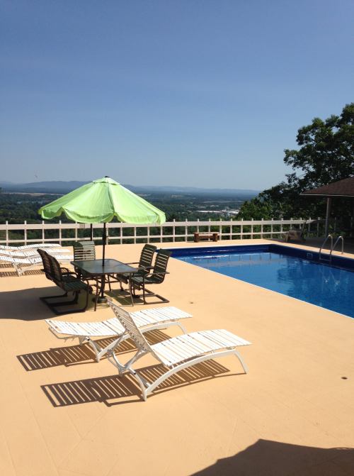 A sunny pool area with lounge chairs, an umbrella, and a scenic view of hills in the background.