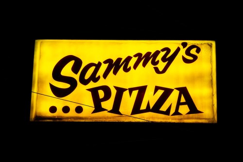 A glowing yellow sign that reads "Sammy's Pizza" in bold, dark letters.