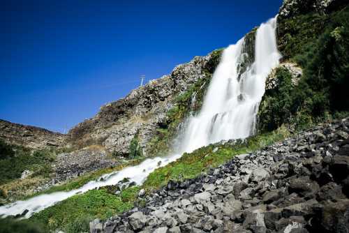 A cascading waterfall flows down rocky terrain, surrounded by lush greenery under a clear blue sky.