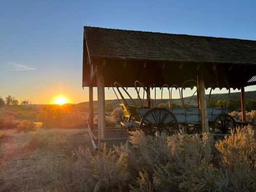 A rustic wagon shelter at sunset, surrounded by sagebrush and rolling hills under a clear sky.