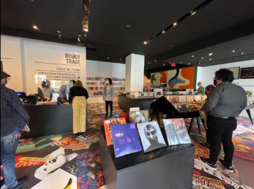 A record store interior with customers browsing vinyl, colorful artwork on the walls, and a vibrant patterned floor.