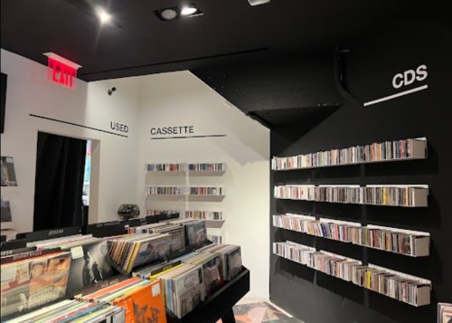 A music store interior featuring shelves of CDs and cassettes, with signs for "Used" and "CDs" on the walls.