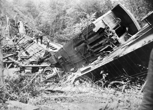 A train wreckage lies on its side amidst trees and debris, with a person inspecting the scene.