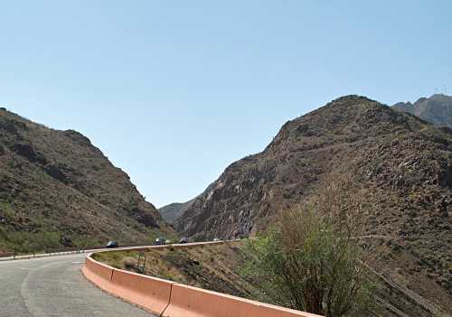 A winding road through rocky hills under a clear blue sky, with sparse vegetation along the roadside.