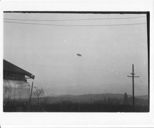 A black and white photo of a rural landscape with a mysterious object in the sky, near a building and power lines.