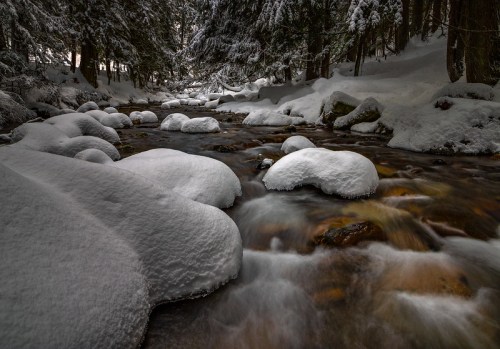 A serene winter scene of a flowing river surrounded by snow-covered rocks and trees.