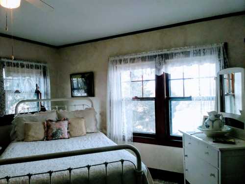 Cozy bedroom with a white metal bed, lace curtains, and a dresser by the window, creating a warm, inviting atmosphere.