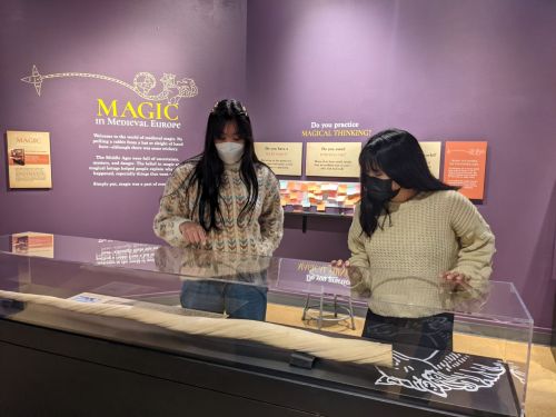 Two women in masks examine a display at a museum exhibit titled "Magic in Medieval Europe."
