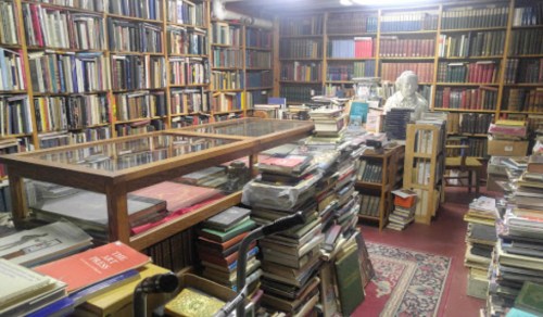 A cozy, cluttered library filled with bookshelves, stacks of books, and a bust statue in a warm, inviting space.