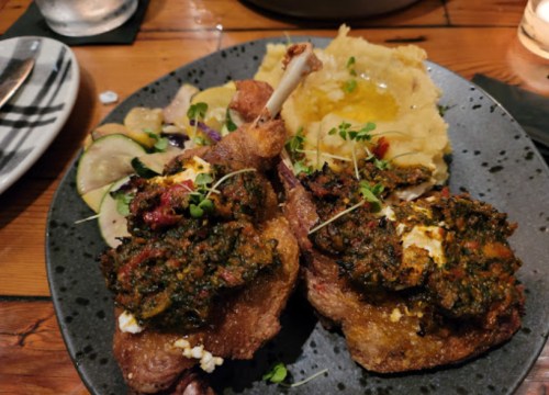 A plate featuring two pieces of meat topped with green sauce, served with mashed potatoes and a side salad.
