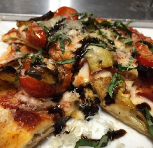 Close-up of a slice of pizza topped with tomatoes, artichokes, cheese, and fresh basil drizzled with balsamic glaze.