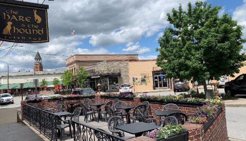 Outdoor seating area of a pub with flower beds, a sign for "The Hare & The Hound," and shops in the background.