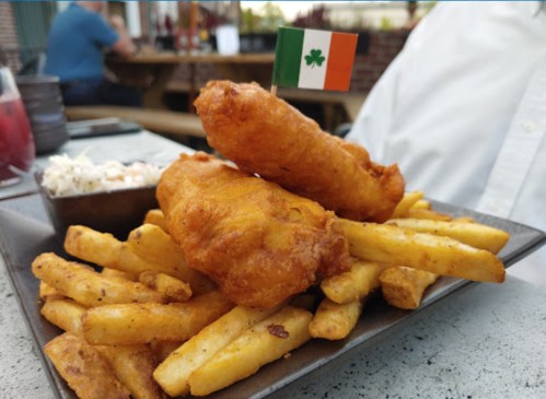 A plate of golden-brown fish and chips, topped with a small Irish flag, alongside a side of coleslaw.