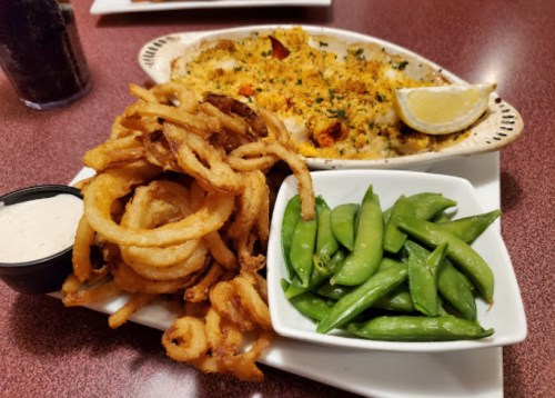 A plate featuring baked seafood with breadcrumbs, curly fries, and fresh green snap peas, garnished with a lemon wedge.
