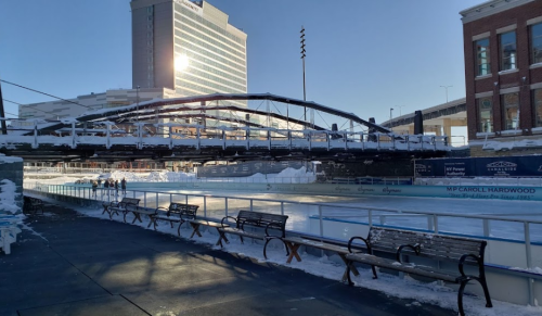 A frozen ice rink under a bridge, with benches nearby and buildings in the background, illuminated by sunlight.