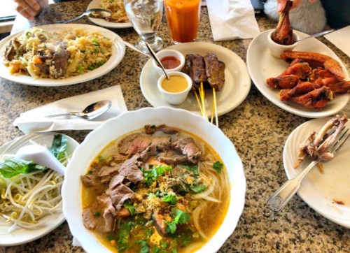 A table spread with various dishes: noodle soup, fried rice, grilled skewers, chicken wings, and dipping sauces.