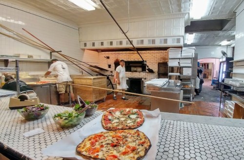 A pizza kitchen with two pizzas in the foreground and chefs preparing food in the background.