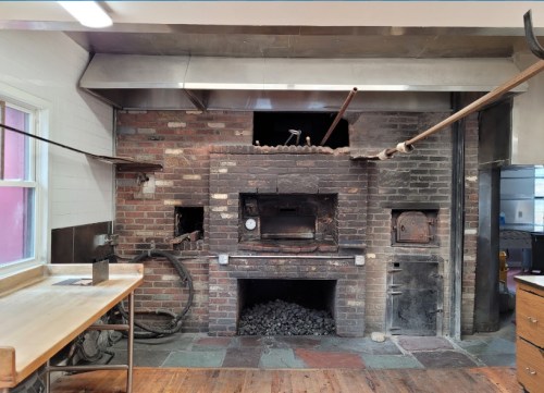 A rustic kitchen with a brick oven, metal accents, and wooden countertops, featuring a large fireplace and cooking tools.