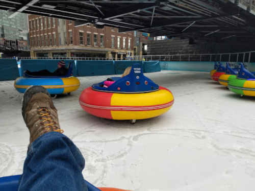 A person in a boot relaxes in a colorful bumper car on an ice rink, with others nearby under a bridge.