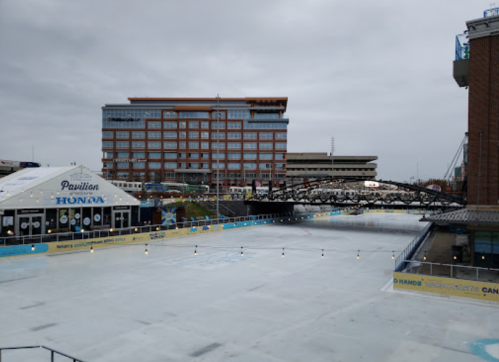 A view of a frozen rink with a building and bridge in the background, under a cloudy sky.