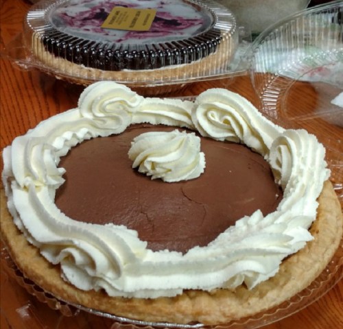 A chocolate cream pie topped with swirls of whipped cream, placed on a wooden table with another pie in the background.