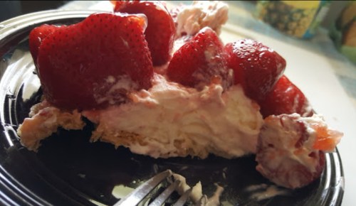 A slice of strawberry dessert with whipped cream and fresh strawberries on a black plate, partially eaten with a fork.
