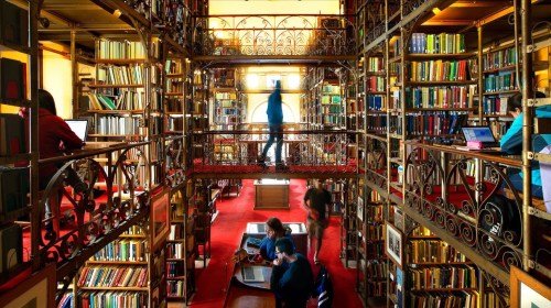 A vibrant library with multiple levels, filled with books and people studying at tables and using laptops.