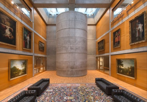 Modern art gallery interior featuring a central concrete column, wooden walls, and paintings on display.