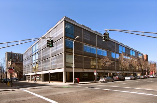 Modern building with large windows at a street intersection, clear blue sky above. Traffic lights visible.