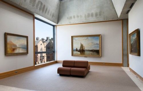 A modern art gallery featuring two large paintings and a brown sofa, with a view of buildings outside.