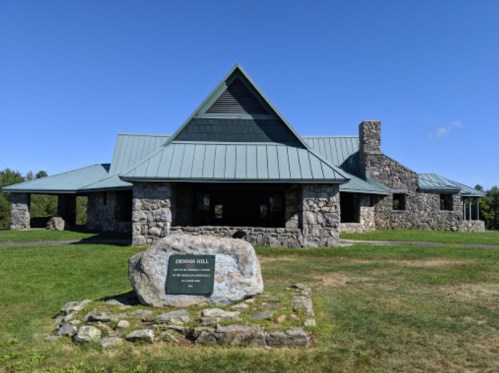 A stone building with a metal roof, surrounded by grass, featuring a sign that reads "Denson Hill." Clear blue sky above.
