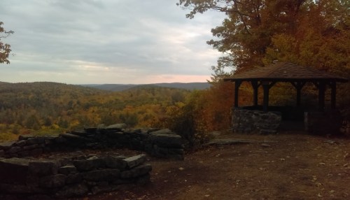 Scenic view of a forested landscape in autumn, featuring a stone gazebo and colorful foliage under a cloudy sky.