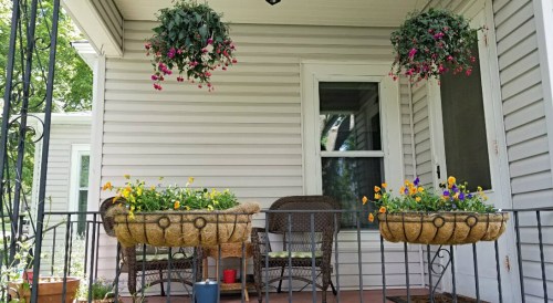 A cozy porch with hanging flower baskets and two planters, featuring colorful flowers and wicker furniture.