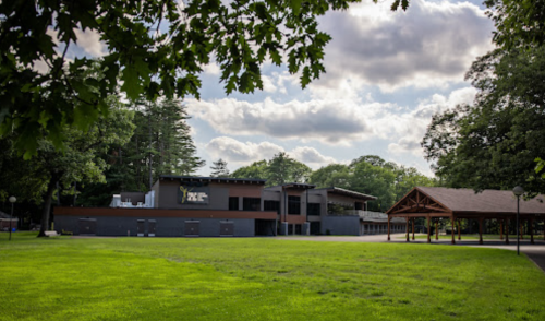 A modern building surrounded by green grass and trees, with a pavilion nearby under a partly cloudy sky.