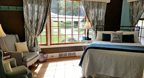 Cozy bedroom with a large window, blue curtains, a bed with white linens, and a comfortable armchair.
