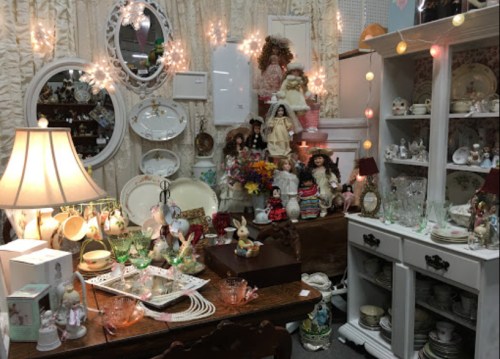 A cozy vintage shop display with dolls, glassware, and decorative items, illuminated by soft lights.