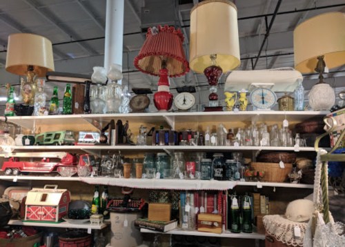 A cluttered shelf filled with vintage items, including lamps, clocks, glass jars, and various collectibles.