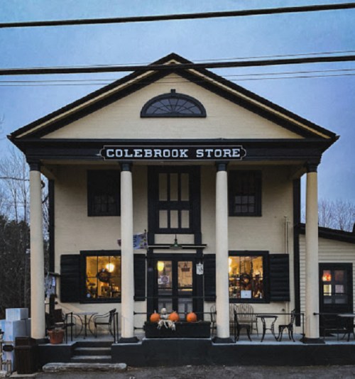 A charming two-story building with columns, labeled "Colebrook Store," featuring pumpkins on the steps and warm lights inside.