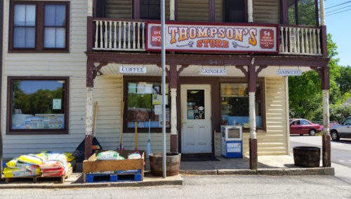 Front view of Thompson's Store, featuring a sign, entrance, and bags of goods outside on a sunny day.