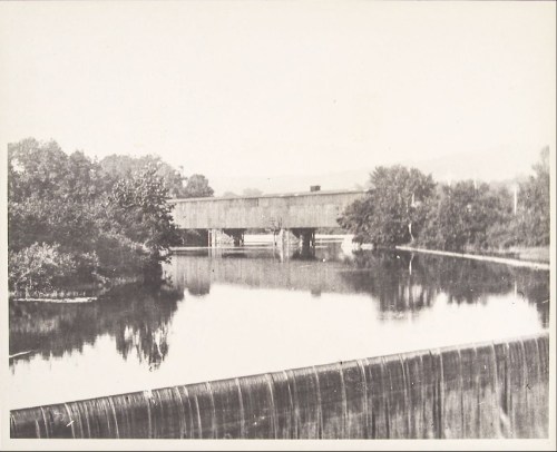 A historic covered bridge spans a calm river, surrounded by trees and distant hills, in a black-and-white photograph.