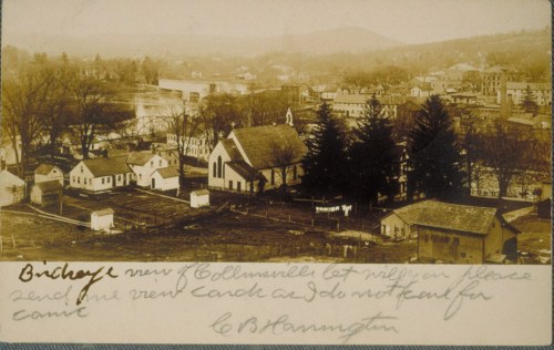 Aerial view of a small town with houses, a church, and trees, featuring a river and mountains in the background.